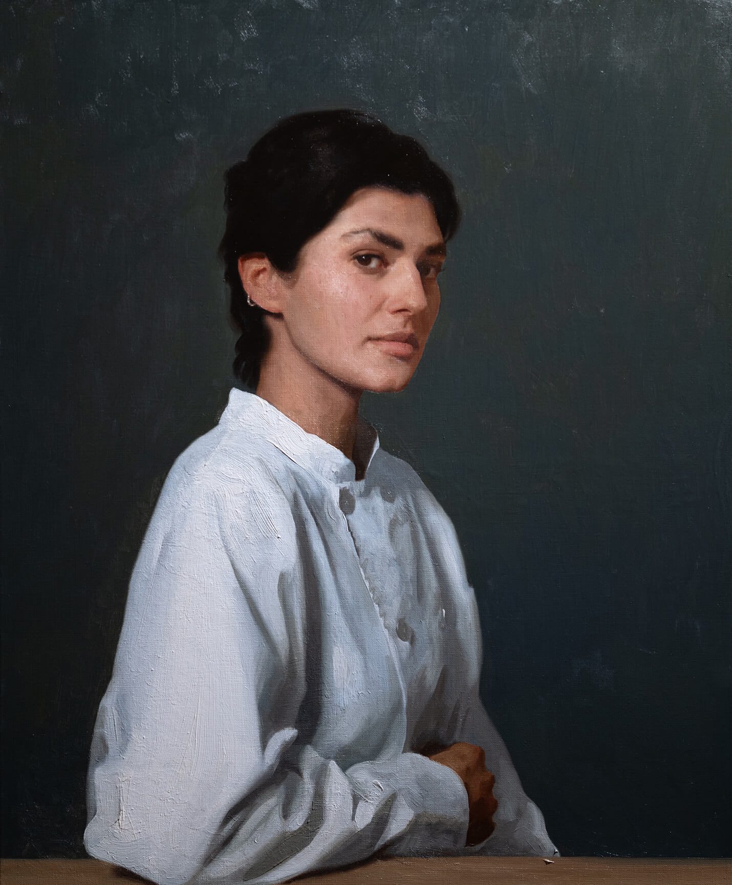Portrait of a woman wearing a chefs uniform, she is sitting leaning with her right hand against a table, you can only see upto her torso, she has dark hair and olive skin and has a pensive look. She sits against a dark grey background