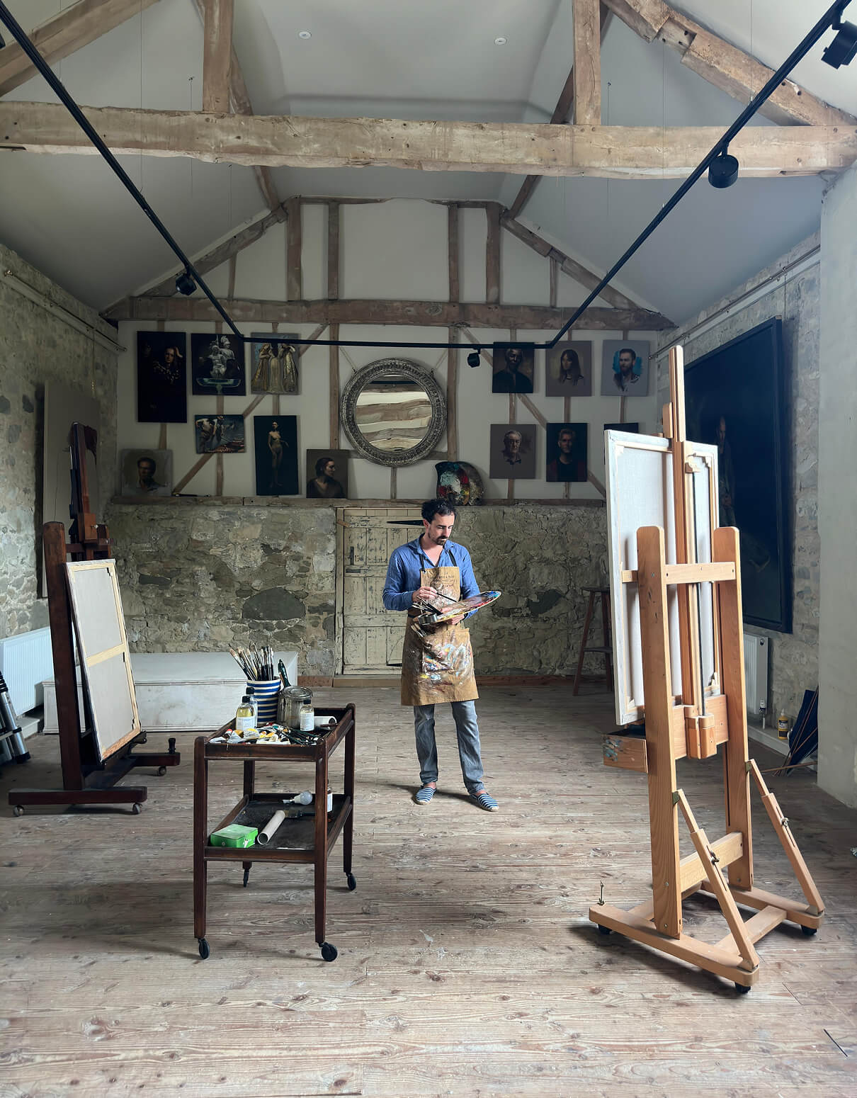 picture of Jamie Coreth painting in his studio. The studio has high ceiling and wooden beams. He is painting a painting which is situated on an easel.
